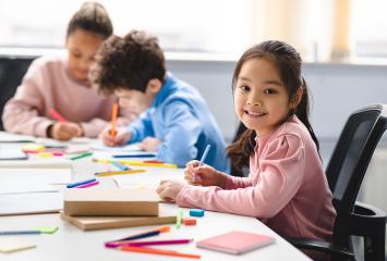 Student smiles at camera, looking up from her drawing, at table surrounded by colorful markers. Two other students focus on their drawings in the background.