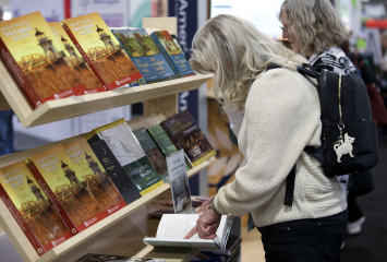 Two women stand in front of a bookshelf. One pages through a book.