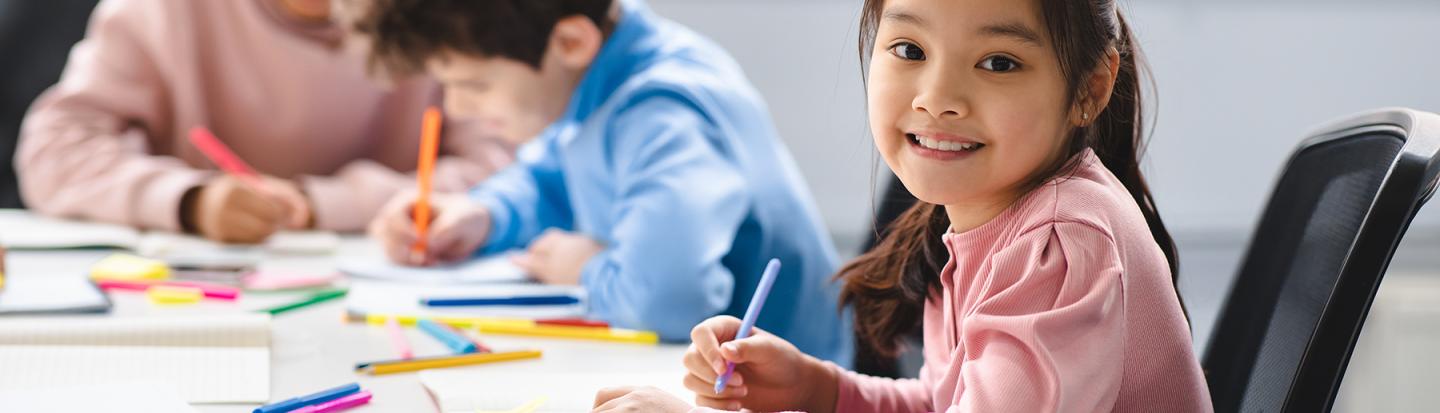 Student smiles at camera, looking up from her drawing, at table surrounded by colorful markers. Two other students focus on their drawings in the background.