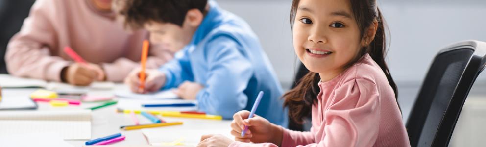 Student smiles at camera, looking up from her drawing, at table surrounded by colorful markers. Two other students focus on their drawings in the background.