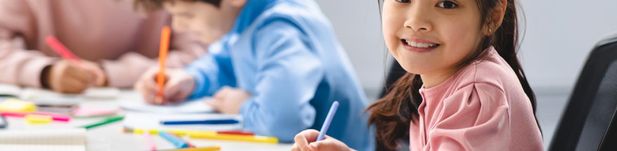 Student smiles at camera, looking up from her drawing, at table surrounded by colorful markers. Two other students focus on their drawings in the background.