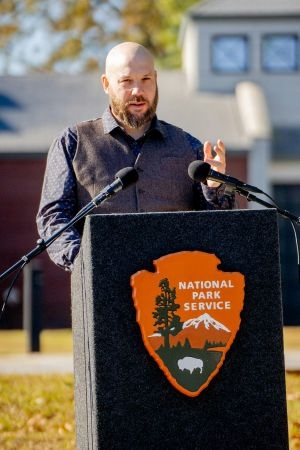 Green and Blue author Daniel Shiels standing at a National Park Service podium outdoors, speaking into two microphones at either side of it.