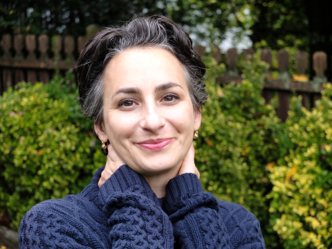 Libby Copeland headshot; a woman with a blue sweater, black and grey hair, and earrings outside in front of shrubs and a fence.